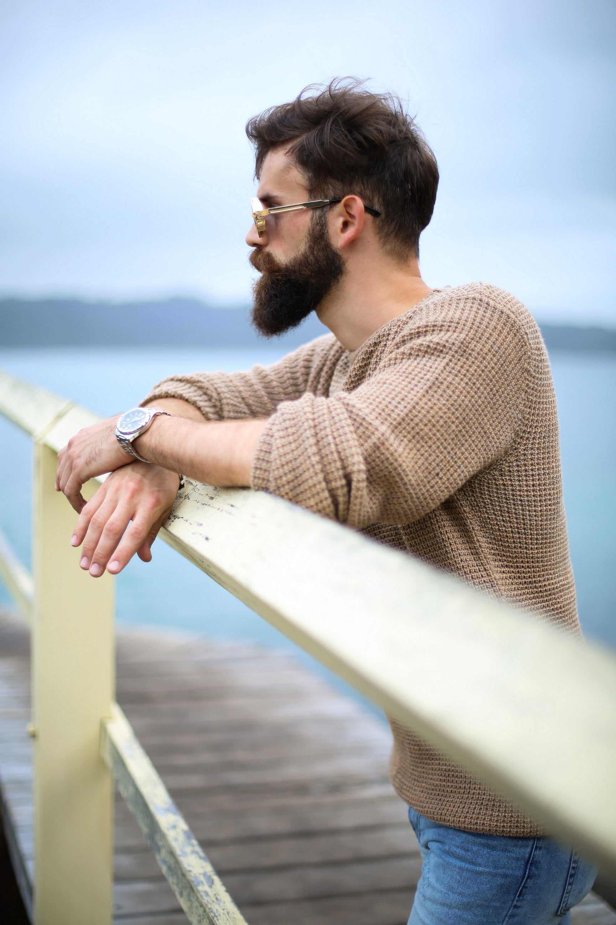 man with thick beard wearing sunglasses and brown sweater leaning on wooden railing outdoors near water